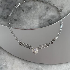 May include: Silver-toned necklace featuring a heart-shaped pendant with a textured design. The necklace has a chain-link style with star-shaped accents. The jewelry is displayed on a reflective surface.