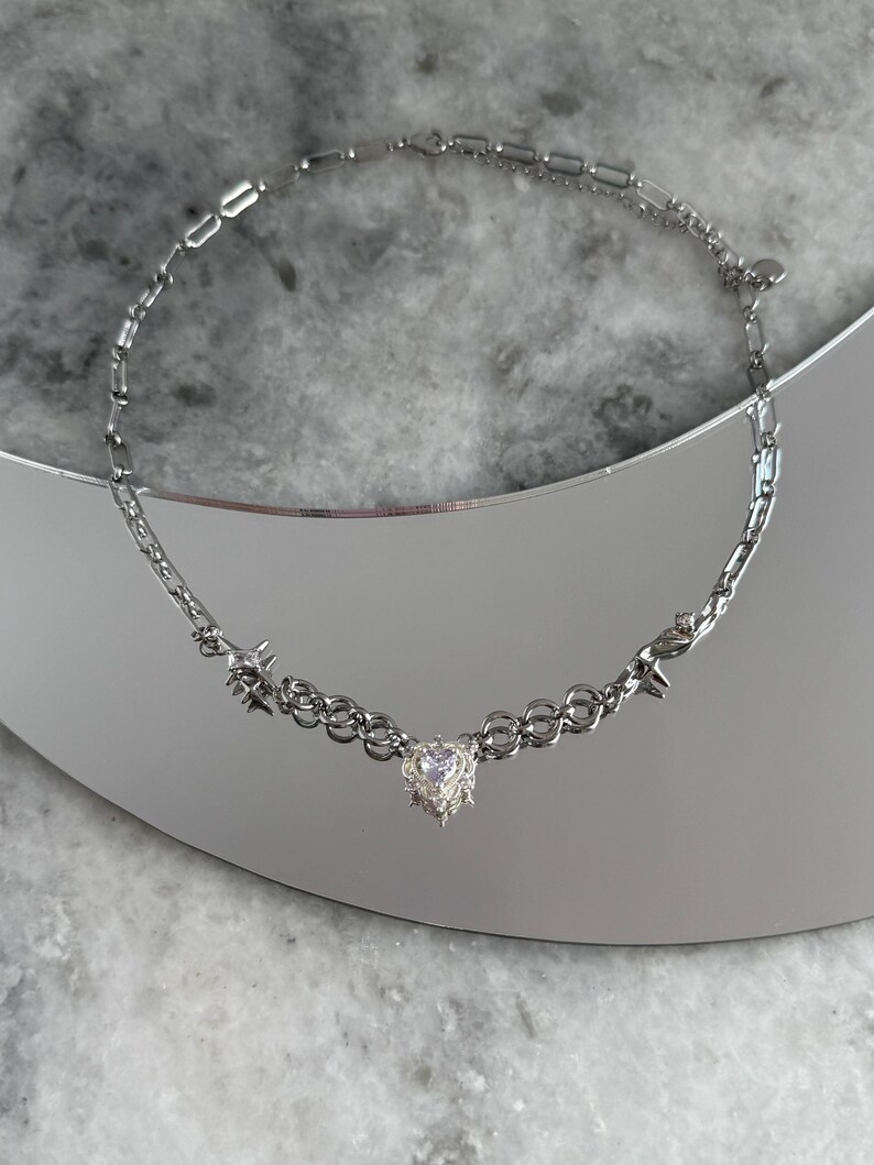 May include: Silver-toned necklace featuring a heart-shaped pendant with a textured design. The necklace has a chain-link style with star-shaped accents. The jewelry is displayed on a reflective surface.