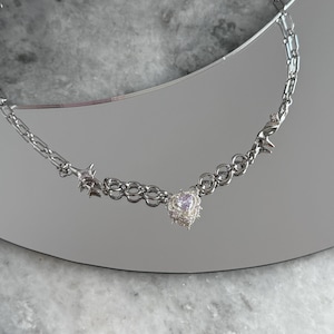 May include: A silver-toned necklace featuring a heart-shaped pendant with a small, light-colored stone. The necklace has a chain design with star and barbed wire accents. The jewelry is displayed on a reflective surface.