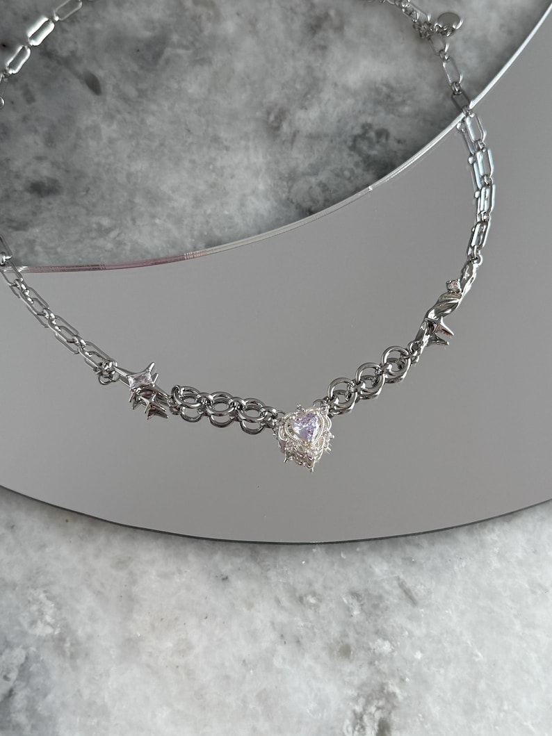 May include: A silver-toned necklace featuring a heart-shaped pendant with a small, light-colored stone. The necklace has a chain design with star and barbed wire accents. The jewelry is displayed on a reflective surface.