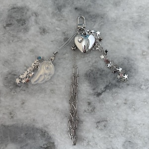Op de afbeelding: Een zilverkleurige sleutelhanger met een hartvormige bedel, een kleine vleugelbedel en stervormige accenten. De sleutelhanger heeft een karabijnhaak en een bungelende ketting.