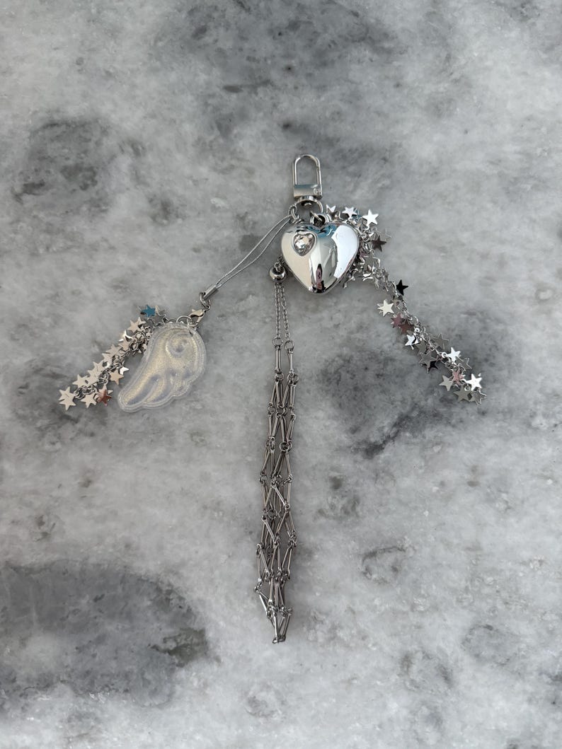 Op de afbeelding: Een zilverkleurige sleutelhanger met een hartvormige bedel, een kleine vleugelbedel en stervormige accenten. De sleutelhanger heeft een karabijnhaak en een bungelende ketting.