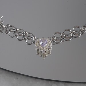 May include: A silver-toned necklace with a heart-shaped pendant. The pendant features a light purple gemstone and ornate detailing. The necklace has a chain link design with star and spike accents. The jewelry is displayed on a reflective surface.