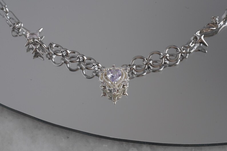 May include: A silver-toned necklace with a heart-shaped pendant. The pendant features a light purple gemstone and ornate detailing. The necklace has a chain link design with star and spike accents. The jewelry is displayed on a reflective surface.