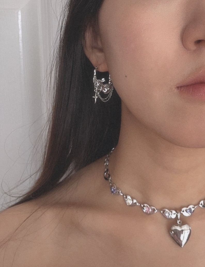Puede incluir: Un collar de plata con un colgante de coraz&oacute;n y una cadena de plata con peque&ntilde;os corazones y piedras de colores. El collar lo lleva una persona con el pelo largo y oscuro y pendientes de plata con un dise&ntilde;o floral.
