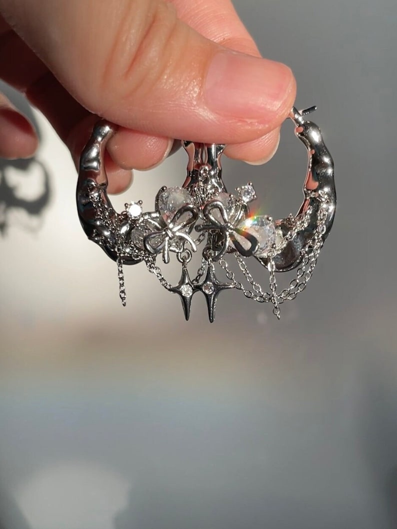 Puede incluir: Pendientes de aro de plata con un dije colgante que presenta una estrella, lazos y cadenas. El dije est&aacute; adornado con cristales transparentes.