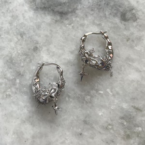 Puede incluir: Un par de pendientes de aro de plata con un dije colgante con forma de lazo y estrella. Los pendientes est&aacute;n hechos de metal texturizado y tienen un dise&ntilde;o delicado y femenino.