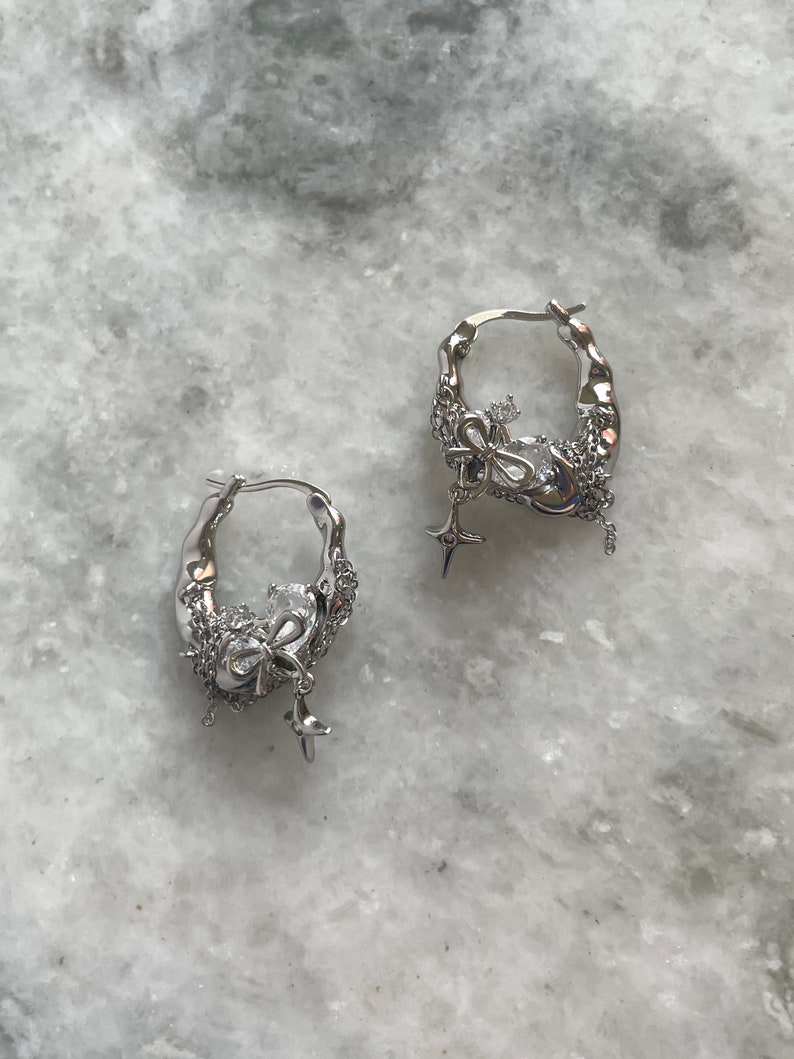 Puede incluir: Un par de pendientes de aro de plata con un dije colgante con forma de lazo y estrella. Los pendientes est&aacute;n hechos de metal texturizado y tienen un dise&ntilde;o delicado y femenino.