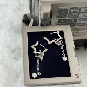 May include: Silver star-shaped earrings with dangling crystal details. The earrings are presented on a dark surface, with a video camera in the background. The earrings feature a delicate design, with a star shape and a dangling crystal.