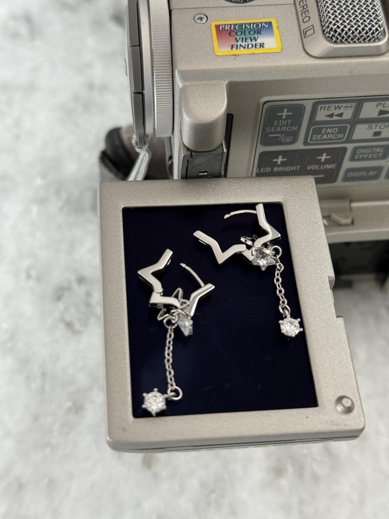 May include: Silver star-shaped earrings with dangling crystal details. The earrings are presented on a dark surface, with a video camera in the background. The earrings feature a delicate design, with a star shape and a dangling crystal.