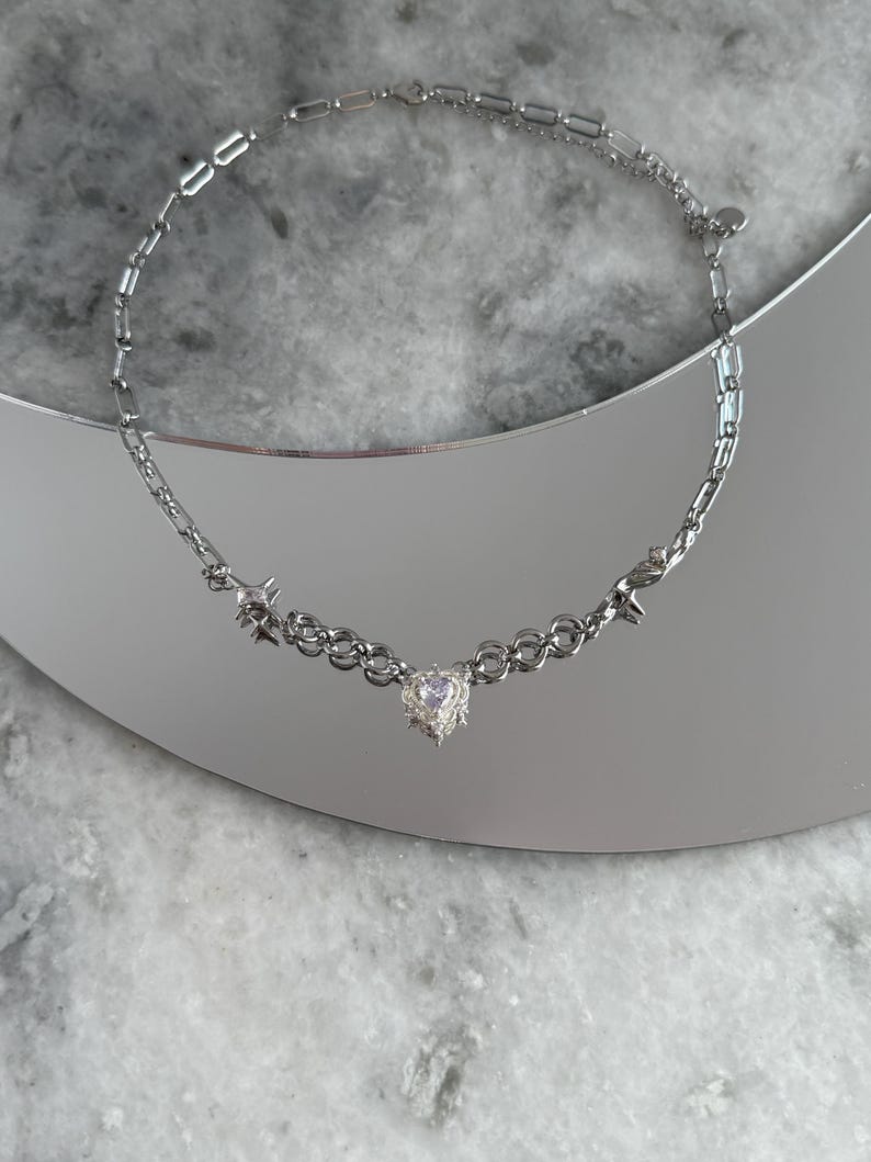 May include: A silver-toned necklace with a heart-shaped pendant and star accents. The necklace features a delicate chain design with a central heart-shaped charm and small star-shaped embellishments. The necklace is displayed on a reflective surface.