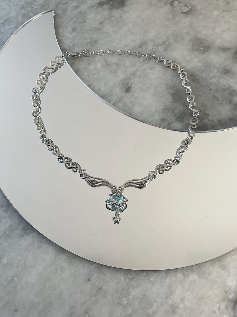 Puede incluir: Un collar de plata con una cadena delicada y adornada y un colgante con una piedra preciosa azul rodeada de piedras m&aacute;s peque&ntilde;as.