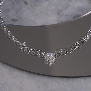 May include: A silver-toned necklace with a heart-shaped pendant. The necklace features a delicate chain design with small star-shaped accents. The pendant is a cluster of small, sparkling crystals. The necklace is displayed on a reflective surface.