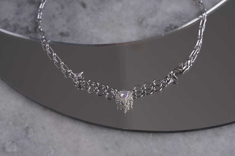 May include: A silver-toned necklace with a heart-shaped pendant. The necklace features a delicate chain design with small star-shaped accents. The pendant is a cluster of small, sparkling crystals. The necklace is displayed on a reflective surface.