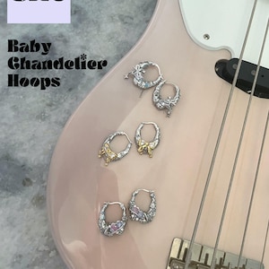 Puede incluir: Primer plano de una guitarra el&eacute;ctrica rosa con cuatro pendientes de aro plateados con abalorios decorativos. Los pendientes est&aacute;n dispuestos en pares en el cuerpo de la guitarra. El texto "Baby Chandelier Hoops" es visible en el cuerpo de la guitarra. El texto "shop more from us" es visible en la parte inferior de la imagen.
