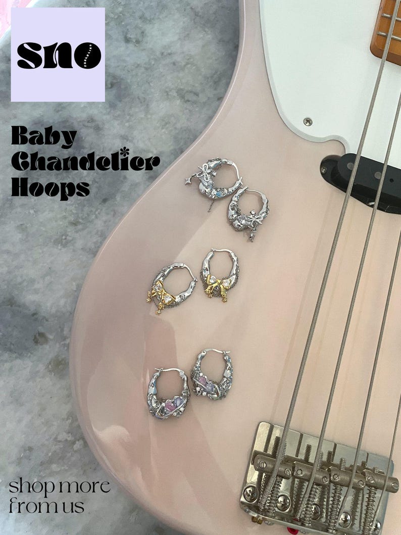 Puede incluir: Primer plano de una guitarra el&eacute;ctrica rosa con cuatro pendientes de aro plateados con abalorios decorativos. Los pendientes est&aacute;n dispuestos en pares en el cuerpo de la guitarra. El texto "Baby Chandelier Hoops" es visible en el cuerpo de la guitarra. El texto "shop more from us" es visible en la parte inferior de la imagen.