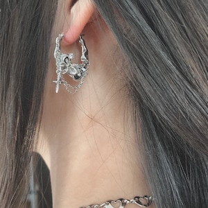 Puede incluir: Pendientes de aro de plata con un dise&ntilde;o detallado que presenta una calavera, una cruz y una cadena. Los pendientes son usados por una persona con cabello largo y oscuro.