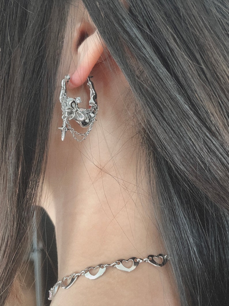 Puede incluir: Pendientes de aro de plata con un dise&ntilde;o detallado que presenta una calavera, una cruz y una cadena. Los pendientes son usados por una persona con cabello largo y oscuro.