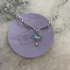 Puede incluir: Un collar de plata con una cadena delicada y un colgante con forma de coraz&oacute;n con una piedra preciosa azul, alas y un dije de estrella.