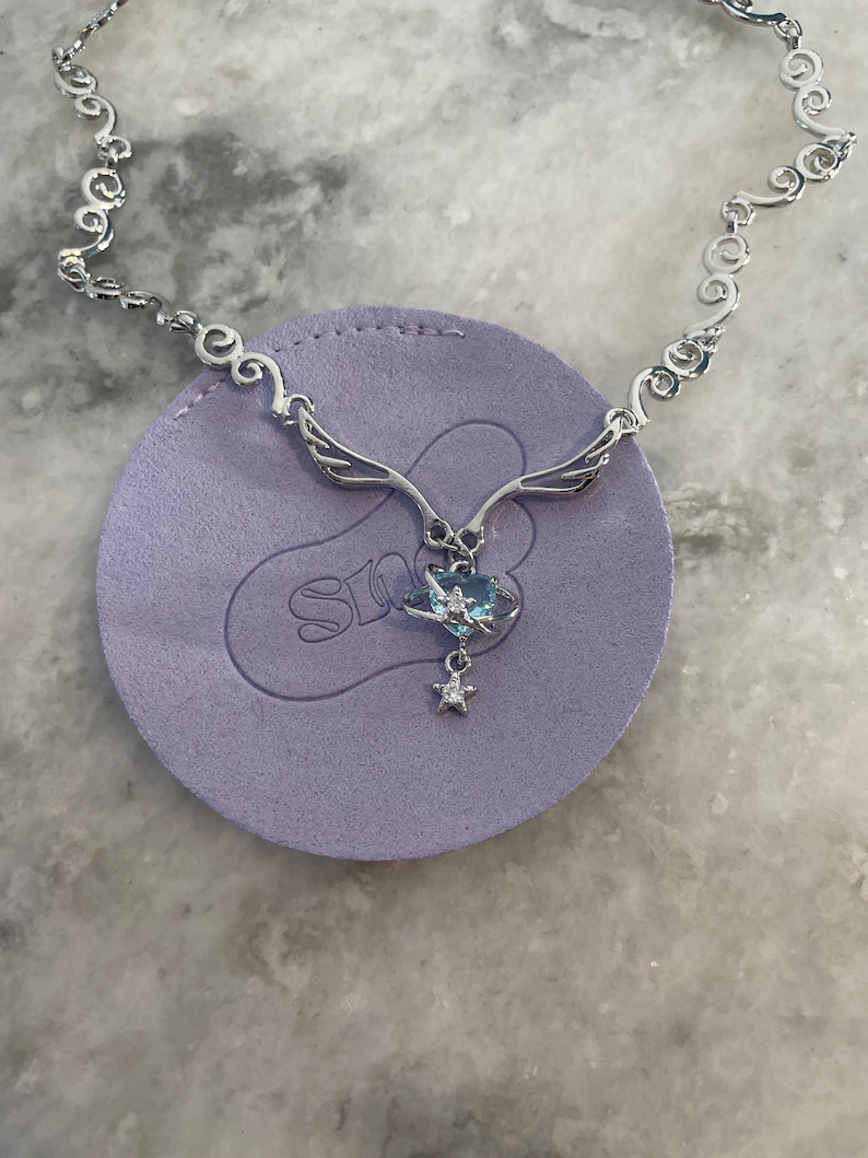 Puede incluir: Un collar de plata con una cadena delicada y un colgante con forma de coraz&oacute;n con una piedra preciosa azul, alas y un dije de estrella.