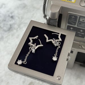 May include: Silver star earrings with dangling chains and clear gemstones. The earrings are presented on a dark blue surface, with a video camera visible in the background.