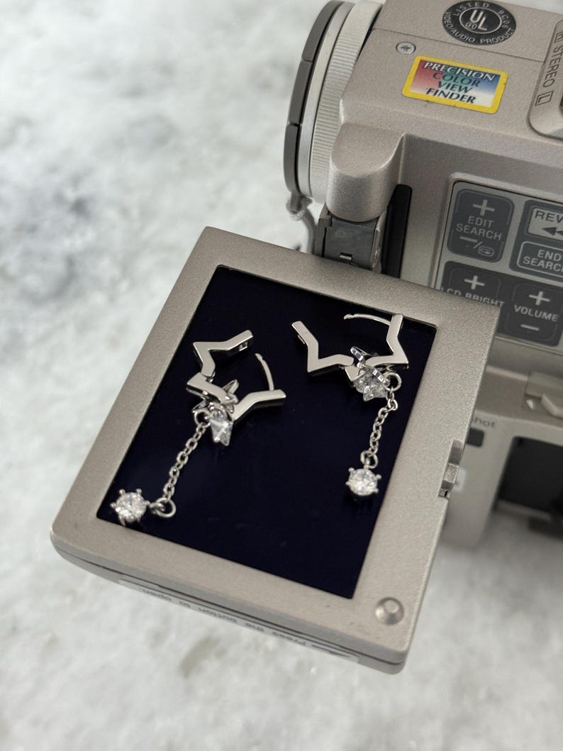 May include: Silver star earrings with dangling chains and clear gemstones. The earrings are presented on a dark blue surface, with a video camera visible in the background.