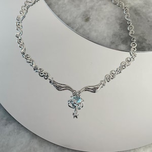 Puede incluir: Un collar de plata con una cadena delicada y adornada y un colgante con una piedra preciosa azul rodeada de piedras m&aacute;s peque&ntilde;as. El colgante tiene forma de estrella con alas.