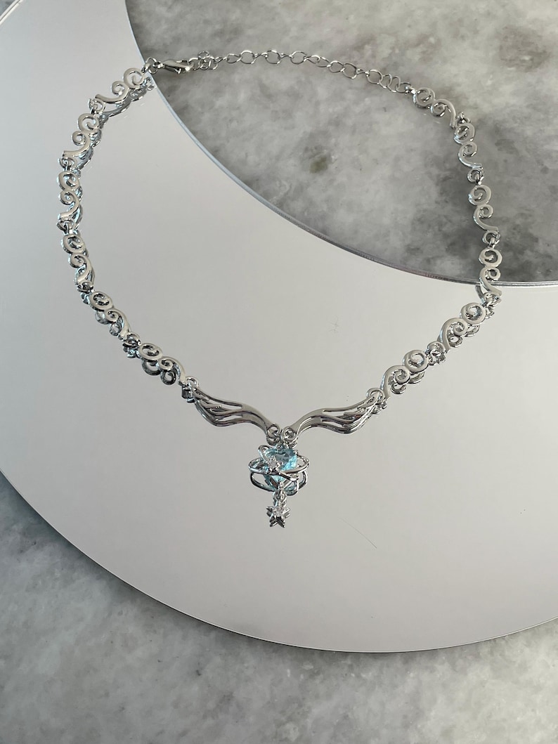 Puede incluir: Un collar de plata con una cadena delicada y adornada y un colgante con una piedra preciosa azul rodeada de piedras m&aacute;s peque&ntilde;as. El colgante tiene forma de estrella con alas.