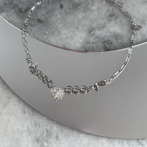 May include: A silver necklace with a heart-shaped pendant featuring a central gemstone. The necklace has a mix of chain styles, including a delicate cable chain and a more substantial link chain. The necklace is displayed on a reflective surface.