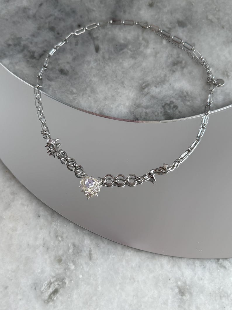 May include: A silver necklace with a heart-shaped pendant featuring a central gemstone. The necklace has a mix of chain styles, including a delicate cable chain and a more substantial link chain. The necklace is displayed on a reflective surface.