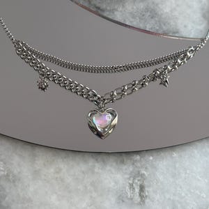 Pode incluir: Um colar prateado em camadas com um pingente em forma de cora&ccedil;&atilde;o. O pingente tem um centro iridescente e opalescente. O colar apresenta uma corrente delicada e uma corrente mais grossa com pingentes de estrela e flor. O colar &eacute; exibido em uma superf&iacute;cie reflexiva.