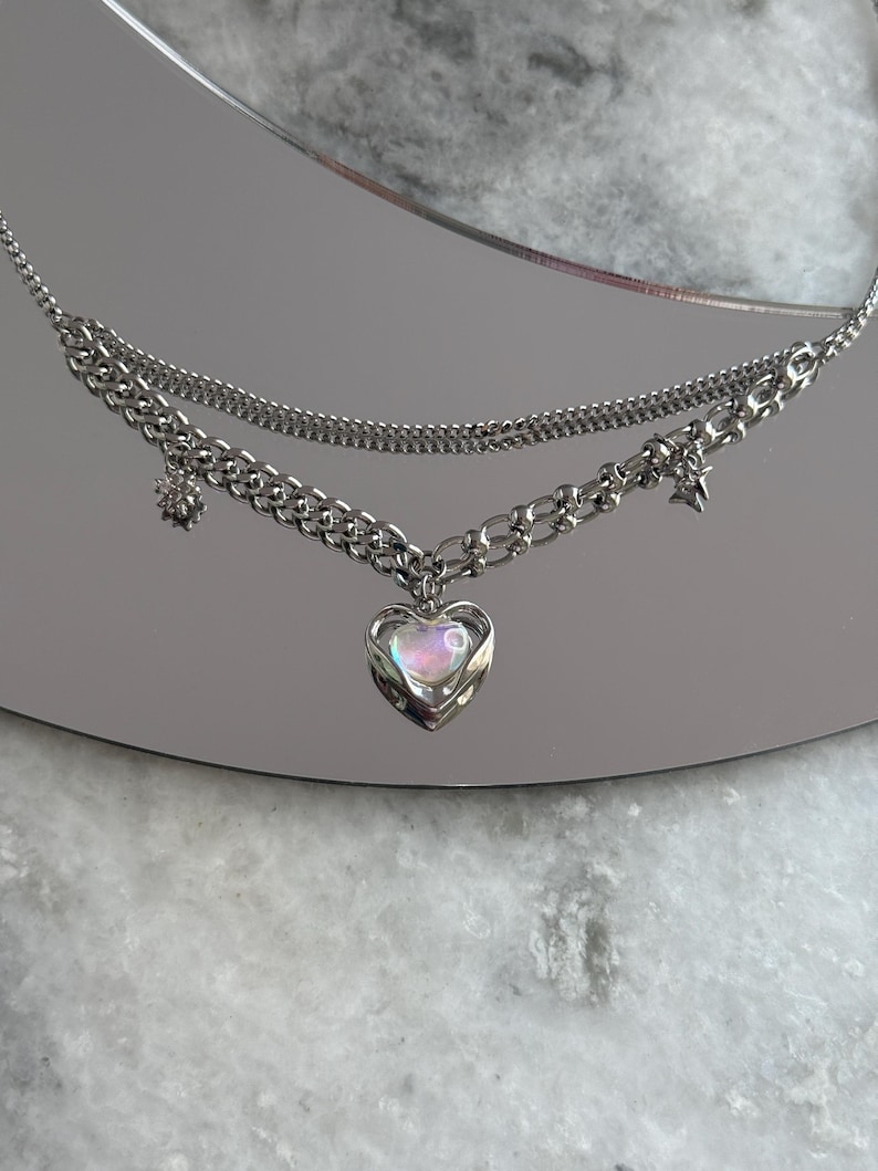 Pode incluir: Um colar prateado em camadas com um pingente em forma de cora&ccedil;&atilde;o. O pingente tem um centro iridescente e opalescente. O colar apresenta uma corrente delicada e uma corrente mais grossa com pingentes de estrela e flor. O colar &eacute; exibido em uma superf&iacute;cie reflexiva.