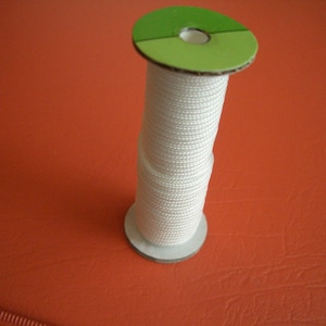 May include: A spool of white cord with a green and white cardboard top and bottom. The cord is tightly wound around the spool. The spool is sitting on an orange surface.