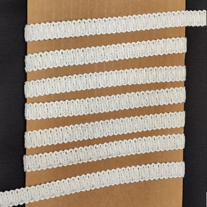 May include: White decorative trim with a scalloped edge, arranged in parallel lines on a brown cardboard surface. The trim has a textured, woven appearance, and is likely used for embellishing clothing or crafts.