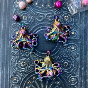 May include: Three iridescent octopus pendants with a rainbow metallic finish are displayed on a dark, textured surface. Scattered around are several iridescent beads and a small glass jar with more beads.