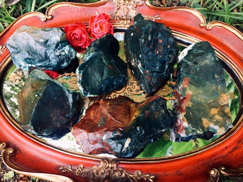 Op de afbeelding: Een verzameling ruwe, natuurlijke stenen in groen, bruin en rood, gerangschikt op een spiegelend oppervlak in een decoratief, sierlijk rood dienblad. Twee rode rozen voegen een vleugje kleur toe.