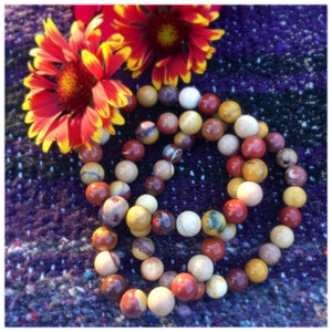 Puede incluir: Una pulsera de cuentas con varias cuentas de tonos tierra, incluyendo colores rojo, marrón, amarillo y crema. Dos flores rojas y amarillas se colocan cerca de la pulsera. Las cuentas tienen aproximadamente 1,3 cm de diámetro.