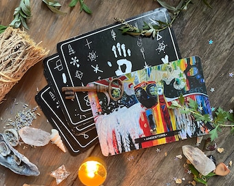 Puede incluir: Un mazo de cartas de tarot en blanco y negro con símbolos, una carta colorida con la palabra "Ancestors" y una llave antigua. Las cartas están dispuestas sobre una superficie de madera con otros elementos espirituales, como una vela y cristales.
