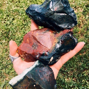 Op de afbeelding: Drie ruwe, ongeslepen edelstenen in tinten donkergroen, rood en zwart worden in een hand gehouden tegen een achtergrond van groen gras. De stenen hebben een ruwe, natuurlijke textuur en verschillende kleurpatronen.
