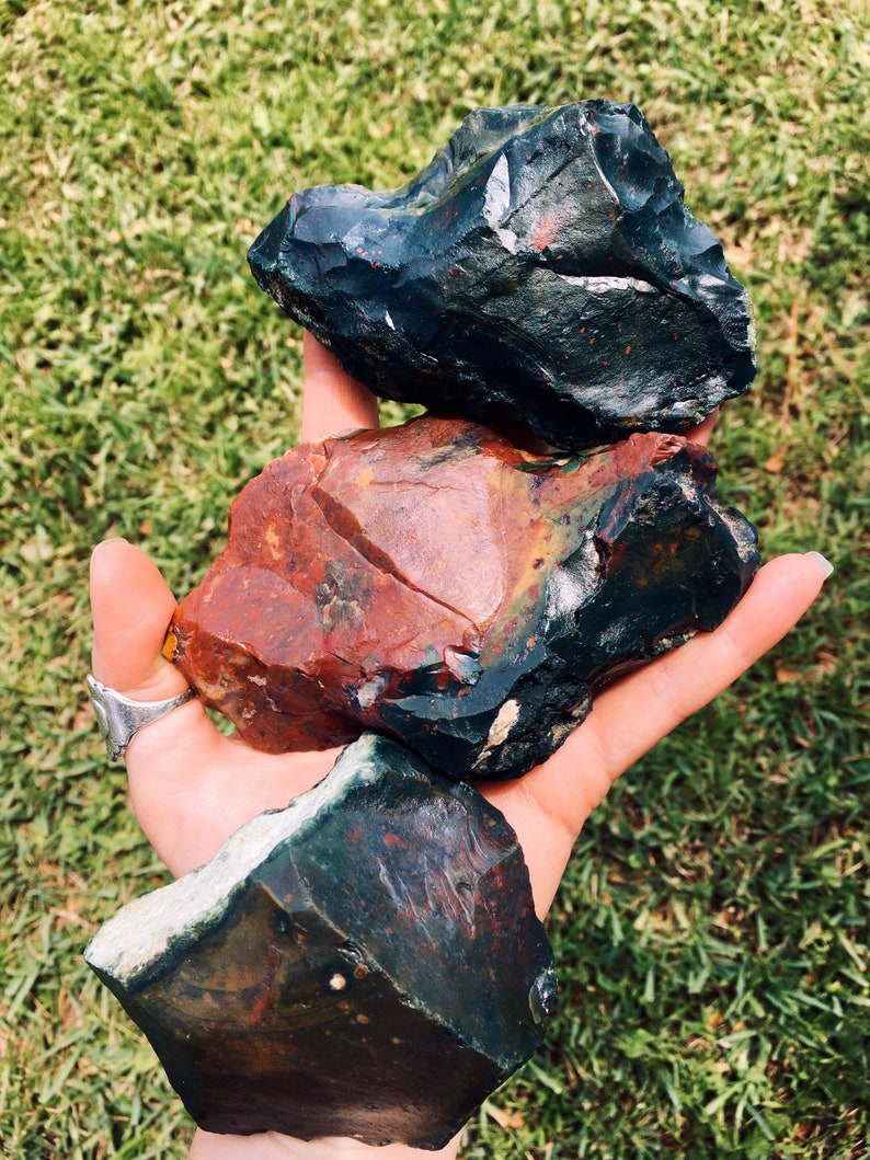 Op de afbeelding: Drie ruwe, ongeslepen edelstenen in tinten donkergroen, rood en zwart worden in een hand gehouden tegen een achtergrond van groen gras. De stenen hebben een ruwe, natuurlijke textuur en verschillende kleurpatronen.