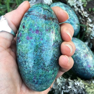 Peut inclure: Une main tient une pierre polie de forme ovale. La pierre a une couleur de base turquoise et verte avec de petites taches irrégulières de rouge foncé. La surface de la pierre est lisse et réfléchissante, avec un aspect naturel et organique. D'autres pierres similaires sont visibles en arrière-plan.