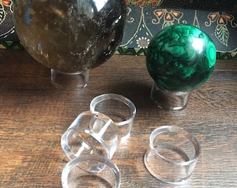 May include: A large smoky quartz crystal ball and a green malachite sphere on clear acrylic stands. Several clear acrylic ring stands are in the foreground. The objects are on a wooden surface.