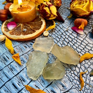 Puede incluir: Un bodegón con cristales translúcidos en bruto, flores secas, una vela encendida y una rodaja de naranja seca. Los elementos están dispuestos sobre una superficie azul texturizada, creando una estética mística y natural.