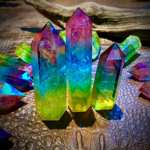 May include: Several colorful quartz crystal points in shades of blue, green, yellow, and purple. The crystals are arranged on a textured, brown surface, with a piece of driftwood in the background. The crystals are polished and have a rainbow effect.