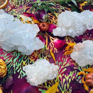 May include: Four clusters of clear quartz crystals are arranged on a colorful, patterned fabric. The crystals are surrounded by dried flowers, rose petals, and small pumpkins. The crystals have a natural, rough texture and varying sizes.