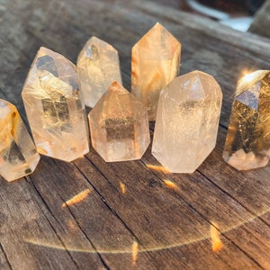 May include: Seven clear quartz crystal points of varying sizes are arranged on a weathered wooden surface. The crystals have a natural, translucent appearance with subtle golden inclusions. The sunlight creates a warm glow and highlights the facets of the crystals.