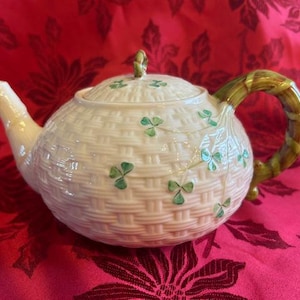 May include: Cream-colored teapot with a woven basket design and green shamrock accents. The handle and lid finial have a bamboo-like appearance. The teapot is set against a red floral background.