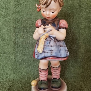 May include: A porcelain figurine of a young girl with blonde hair wearing a plaid dress and pink socks. She is holding knitting needles and yarn in her hands. A small ball of yarn is at her feet.
