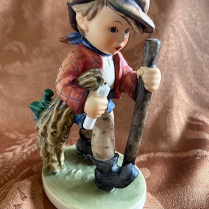 May include: A porcelain figurine of a boy wearing a plaid shirt and cap, holding a stick and a bundle of twigs. He is standing on a green base.