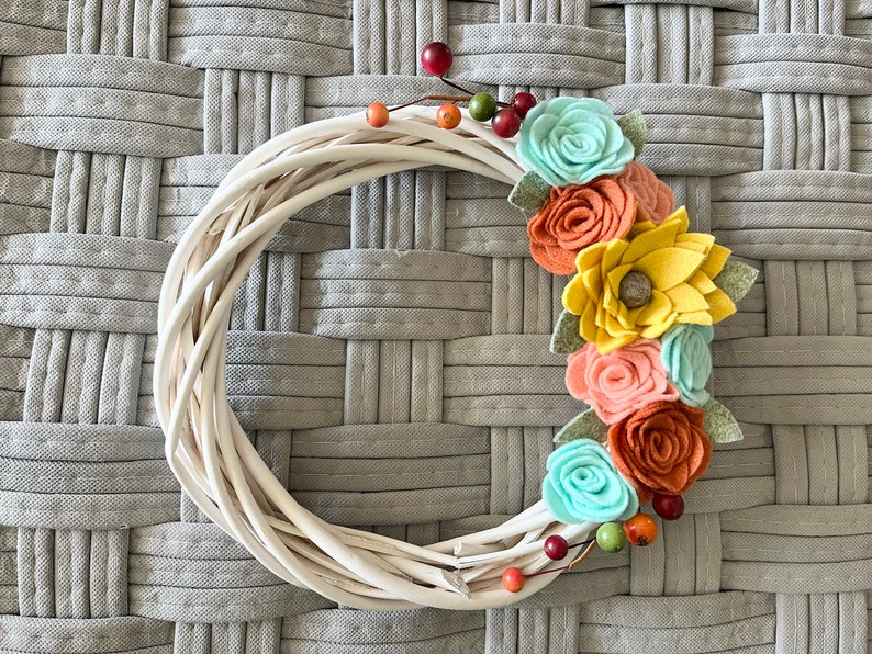 May include: A white twig wreath decorated with felt flowers in shades of yellow, orange, pink, and blue. The flowers are arranged in a cluster on the right side of the wreath.